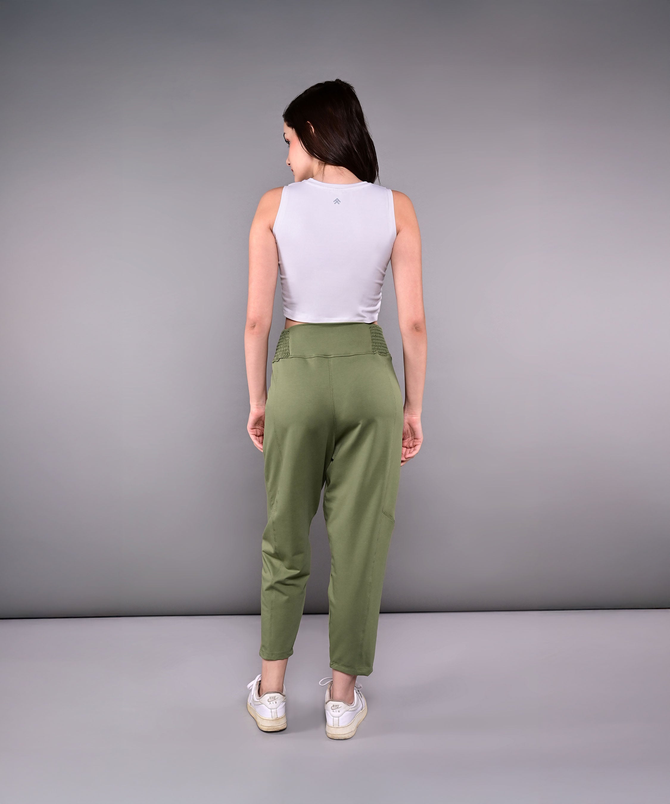 A woman stands facing away against a gray background, wearing the Pleated Yoga Pant With Smocked Waistband in green and a white sleeveless top.