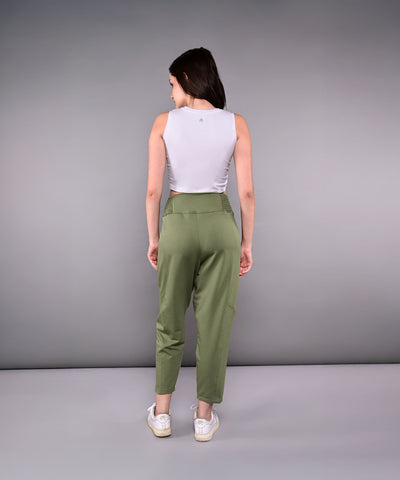 A woman stands facing away against a gray background, wearing the Pleated Yoga Pant With Smocked Waistband in green and a white sleeveless top.