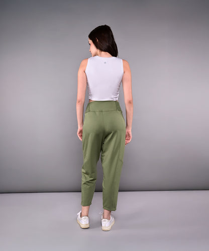A woman stands facing away against a gray background, wearing the Pleated Yoga Pant With Smocked Waistband in green and a white sleeveless top.