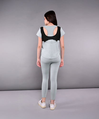 A woman in the Dynamic Grey Performance Set—light gray athletic leggings and a racerback top—faces away, wearing a black posture corrector over her workout outfit.