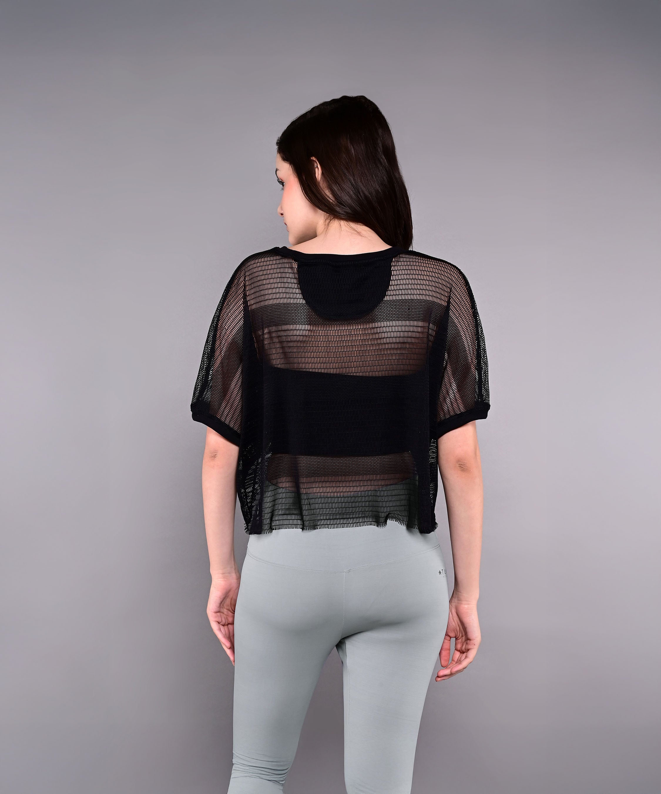 A woman faces away, wearing the Tuna Active Black Netted Top and light gray pants, set against a plain gray background.