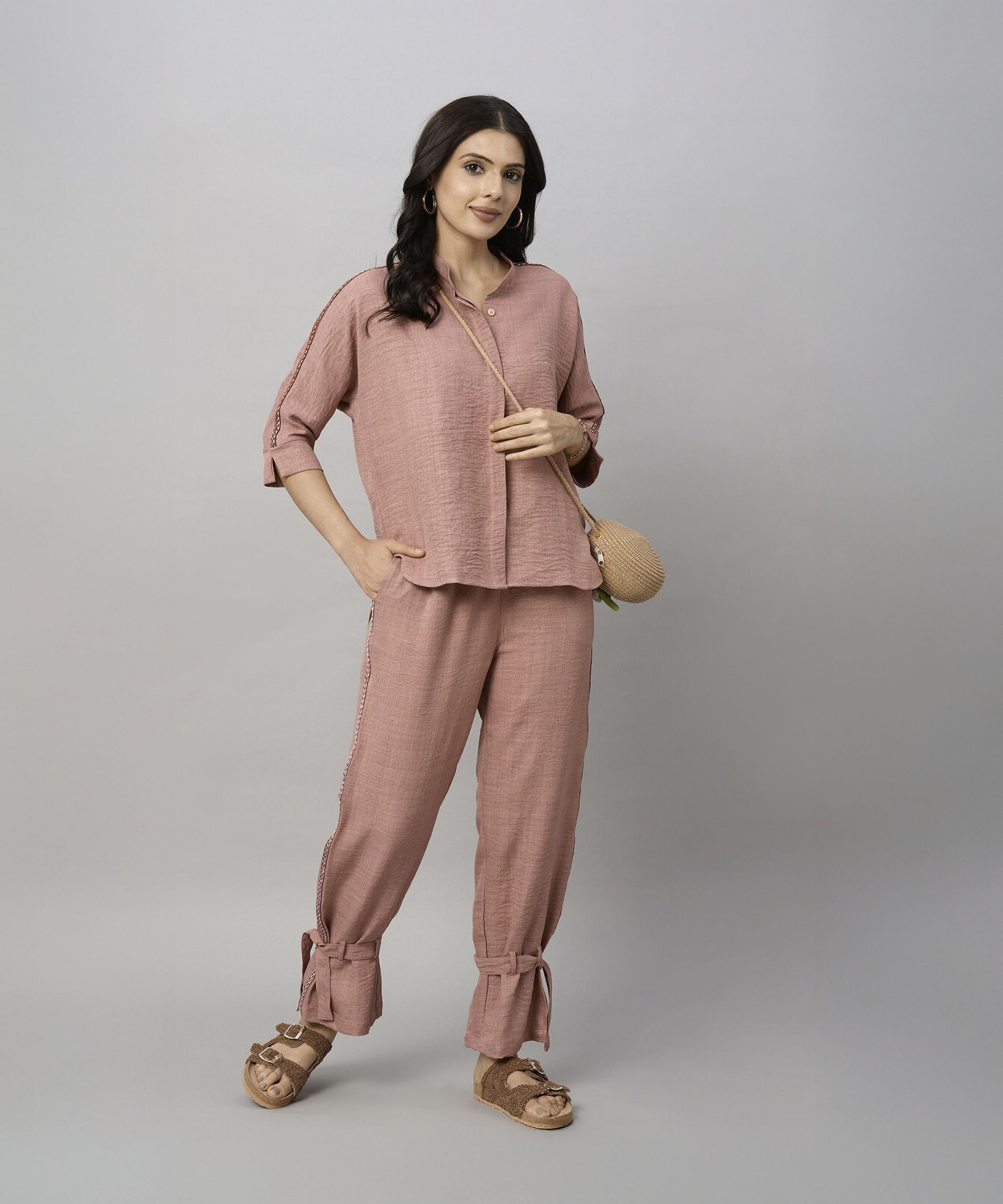 A woman poses against a plain gray background wearing the Vintage Pink Loose Shirt and Tie Up Pant Cord Set, styled with a straw bag and sandals.