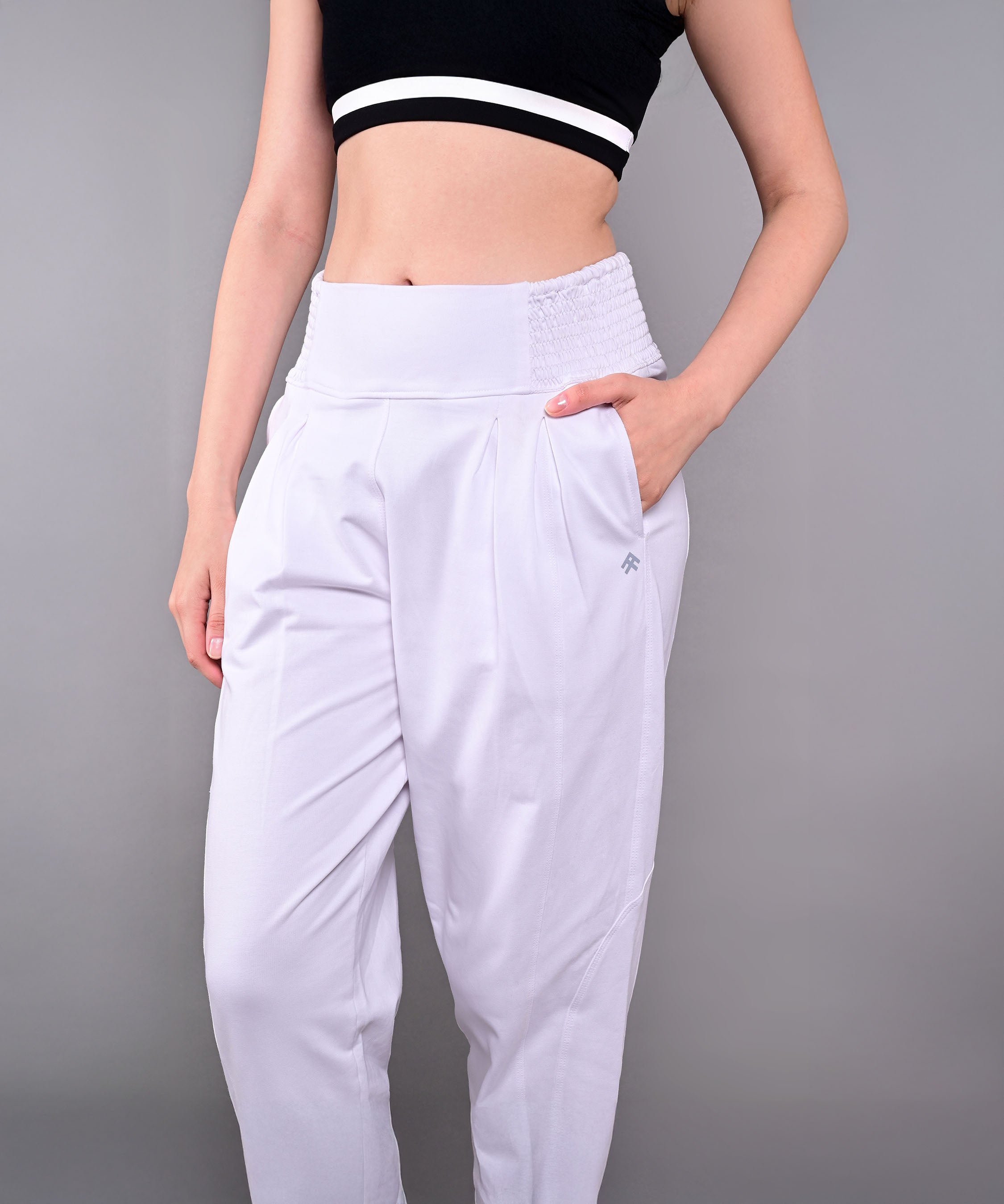 A woman wears Pleated Yoga Pant With Smocked Waistband in white, high-waisted and pleated, paired with a black crop top, standing with one hand in her pocket.