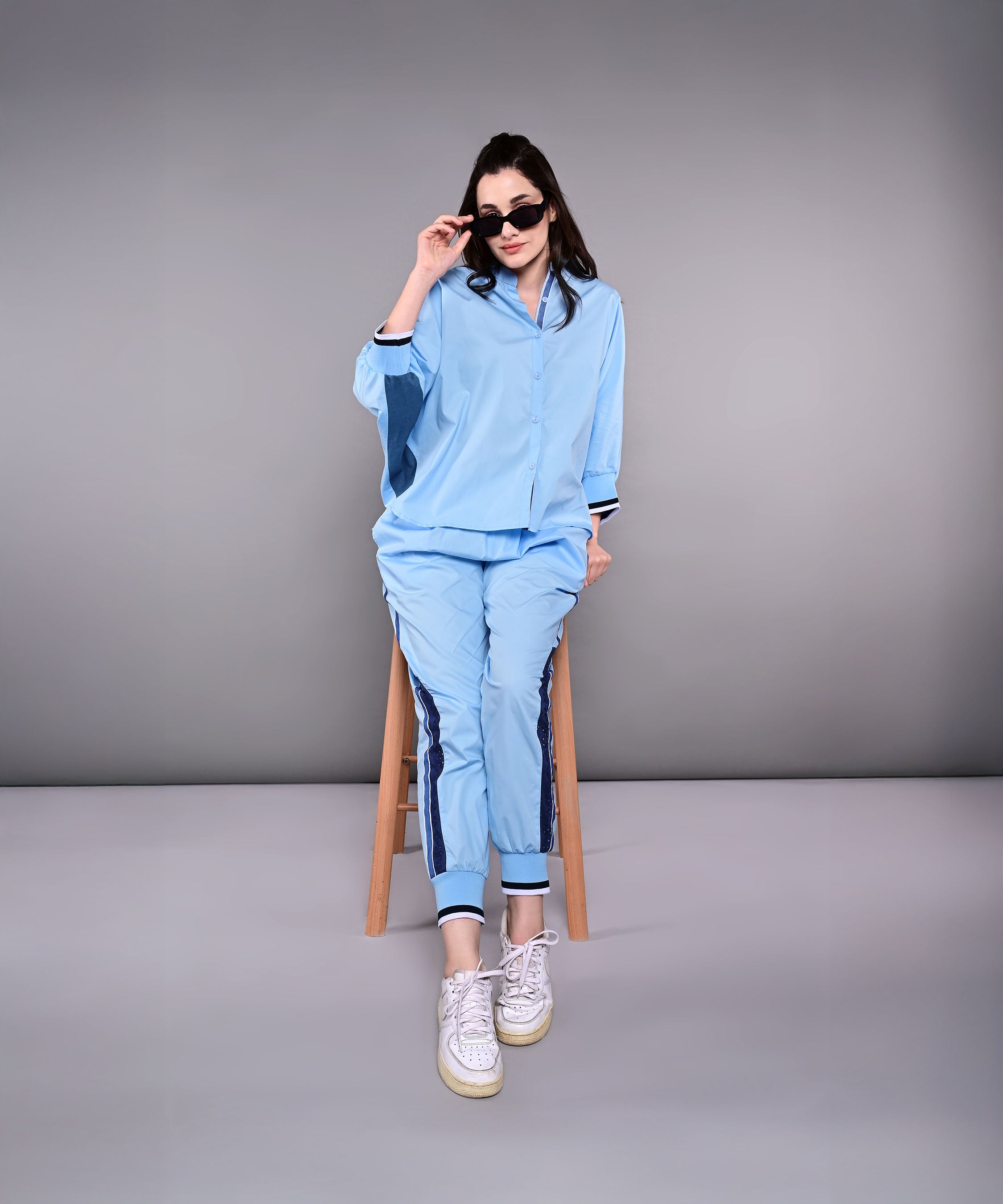 A woman in the Denim Accented Set—100% cotton shirt and joggers—sits on a stool, wearing sunglasses and white sneakers, against a plain gray background.