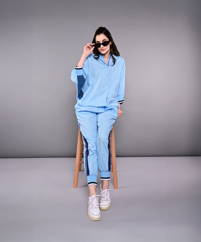 A woman in the Denim Accented Set—100% cotton shirt and joggers—sits on a stool, wearing sunglasses and white sneakers, against a plain gray background.