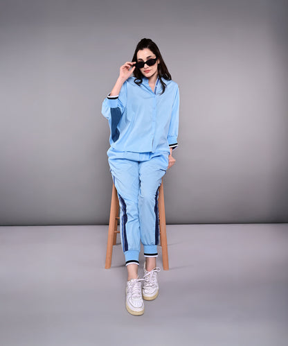 A woman in the Denim Accented Set—100% cotton shirt and joggers—sits on a stool, wearing sunglasses and white sneakers, against a plain gray background.