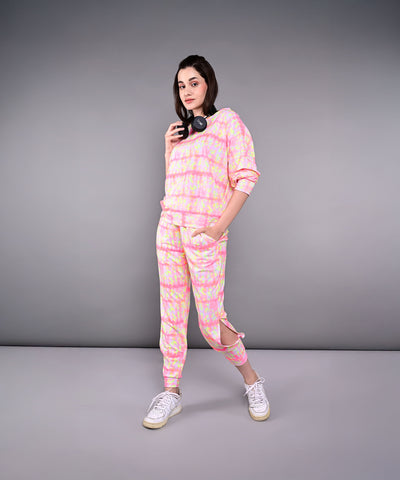 Woman wears the Comfort Fit Tie-Dye Sweatshirt With Joggers Set in pink and yellow, made from breathable poly-blend fabric, paired with white sneakers and headphones, standing against a gray background.