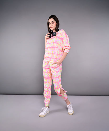 Woman wears the Comfort Fit Tie-Dye Sweatshirt With Joggers Set in pink and yellow, made from breathable poly-blend fabric, paired with white sneakers and headphones, standing against a gray background.