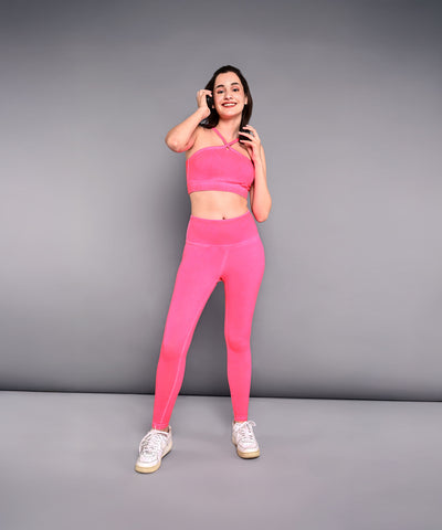 A smiling woman wears the Neon Pink Sports Bra, made from skin-friendly cotton lycra, and poses against a gray wall while holding her hair.