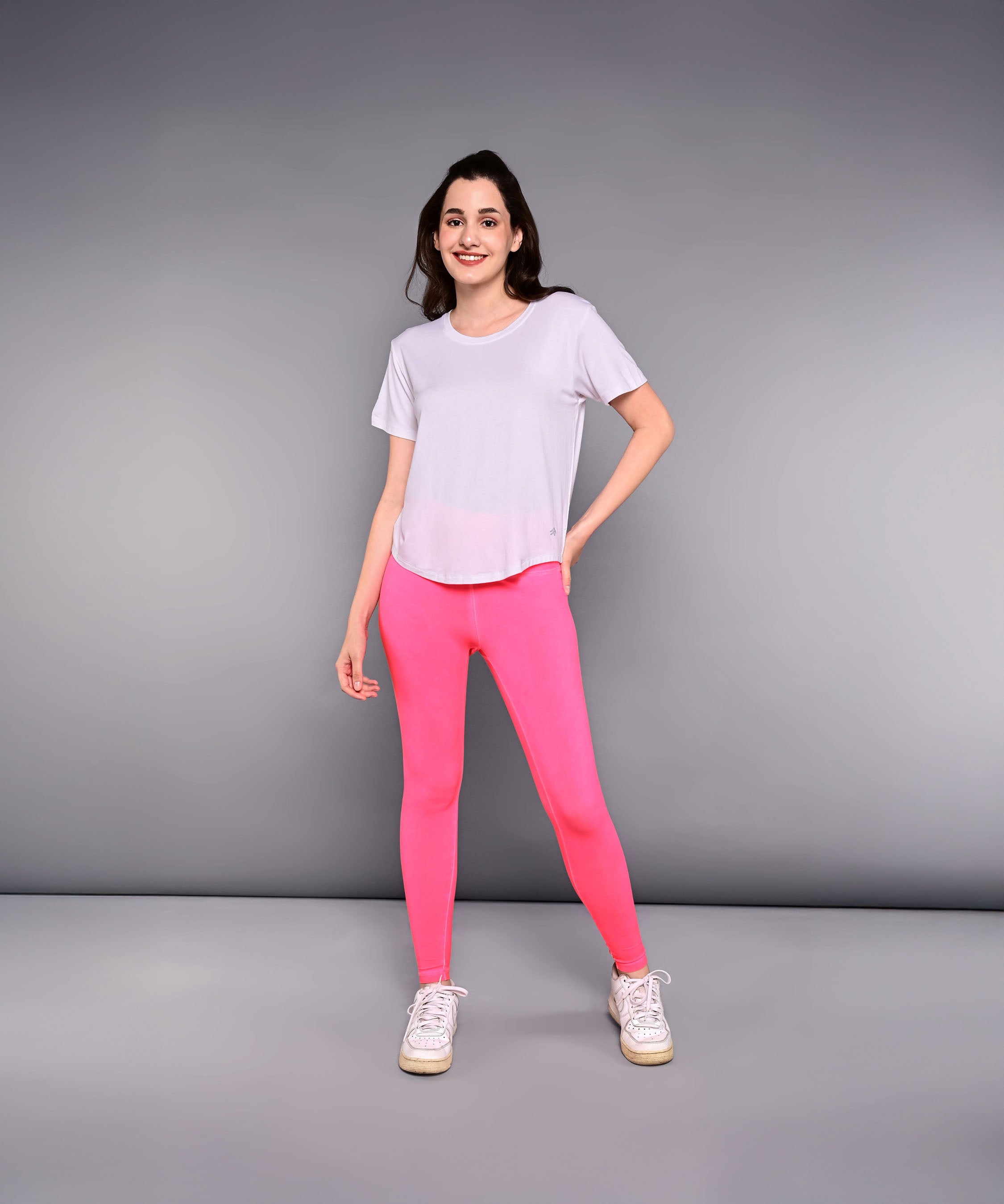 A woman stands against a gray background wearing the 'Pilates Has Your Back' Printed Tee—a white cotton modal shirt—paired with bright pink leggings and white sneakers.