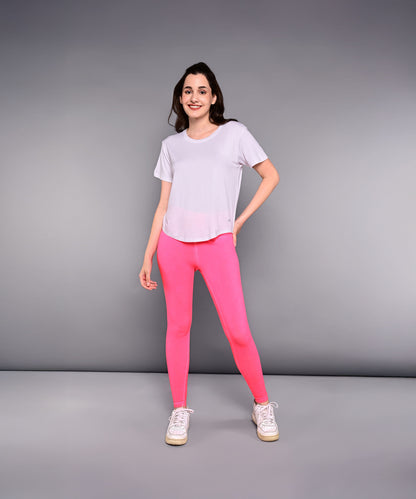 A woman stands against a gray background wearing the 'Pilates Has Your Back' Printed Tee—a white cotton modal shirt—paired with bright pink leggings and white sneakers.