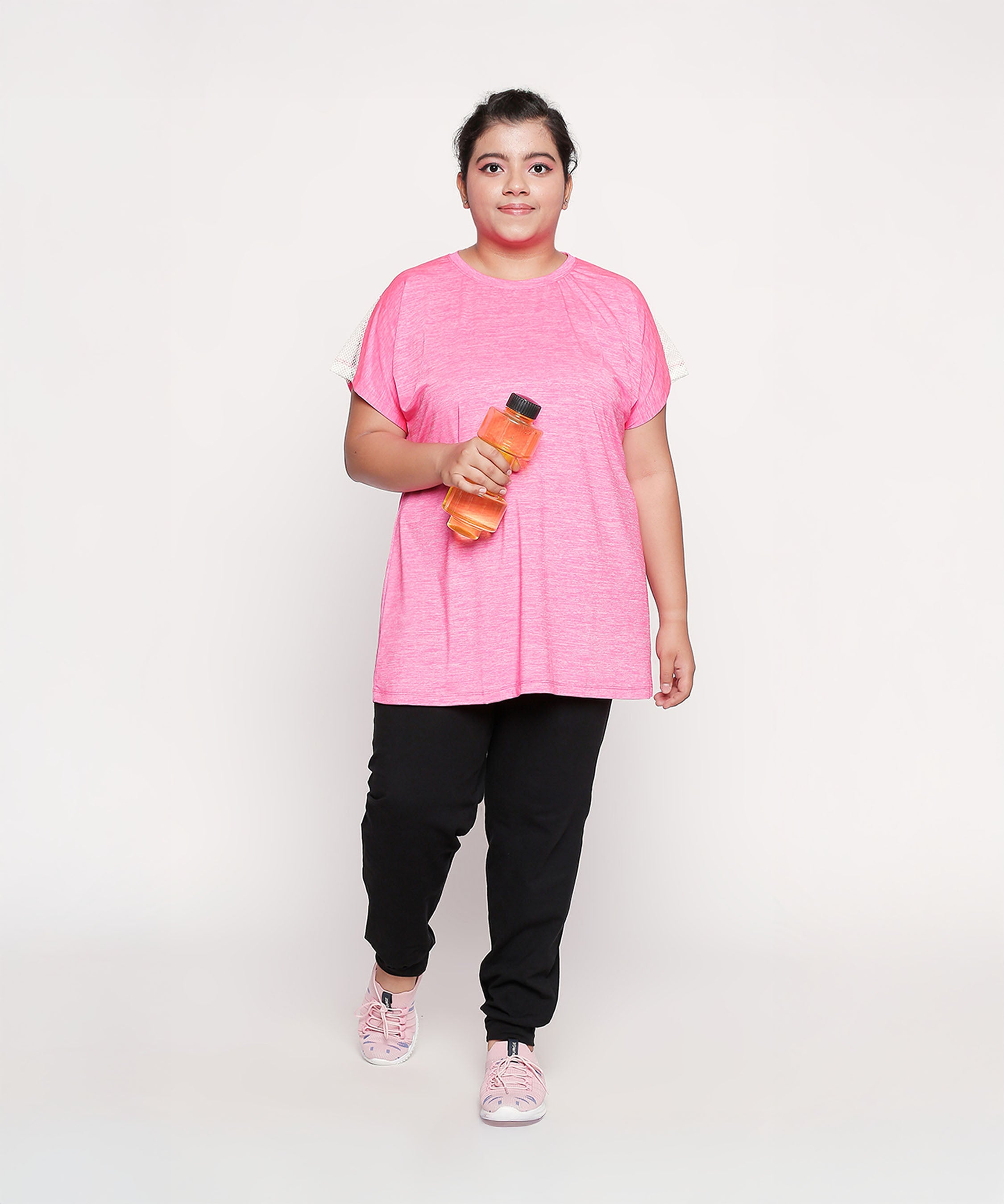 A woman in a pink Nova Sports Tee and black pants holds an orange water bottle, standing against a white background.