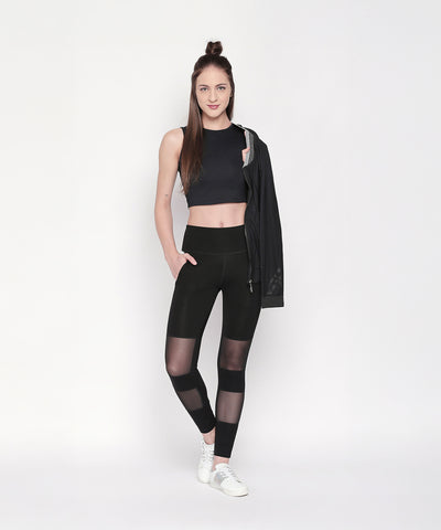 A woman wearing the Active Wear Crop Top in Black Colour, mesh-paneled leggings, and white sneakers, holding a black jacket and standing against a white background.