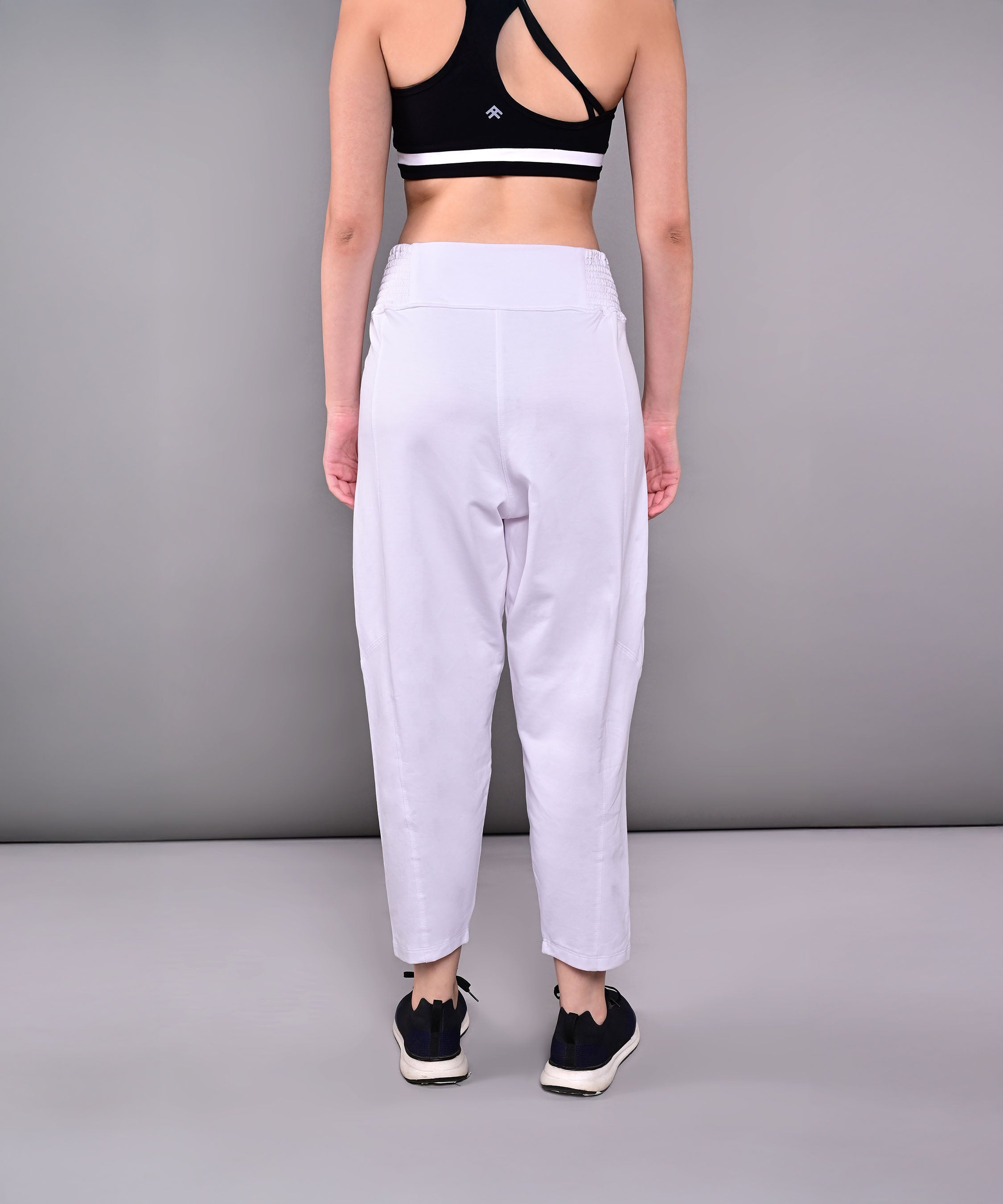 A woman stands with her back to the camera against a gray backdrop, wearing a black sports bra and Pleated Yoga Pant With Smocked Waistband in white cotton.