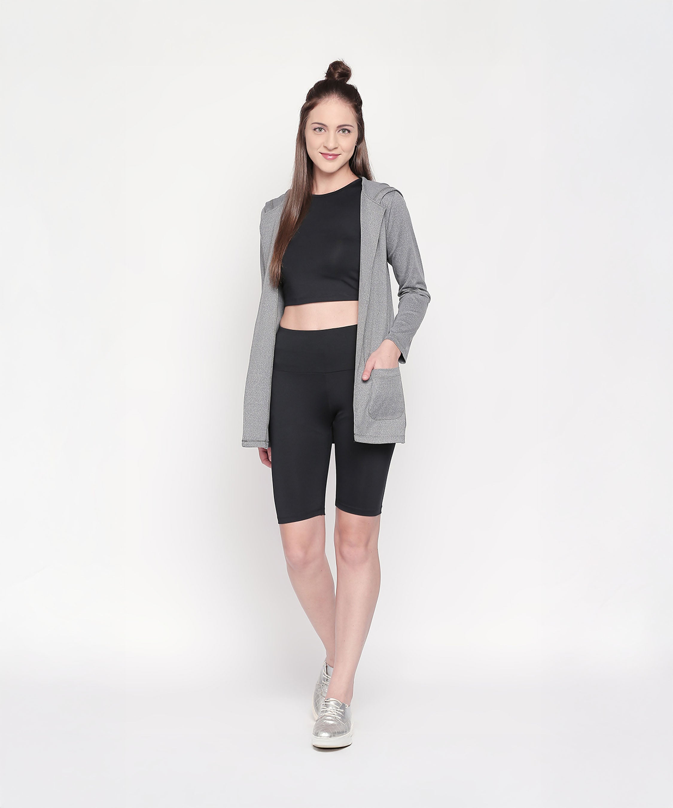 A woman stands and smiles on a white background, wearing a gray hooded jacket, black crop top, and Black Biker/Cycling Shorts featuring a high-rise waistband.