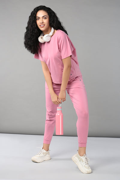 A woman in the Flamingo Pink Chic Ensemble, featuring a racer back design and buttery soft fabric, with headphones around her neck and a pink bottle in hand, stands against a gray background.