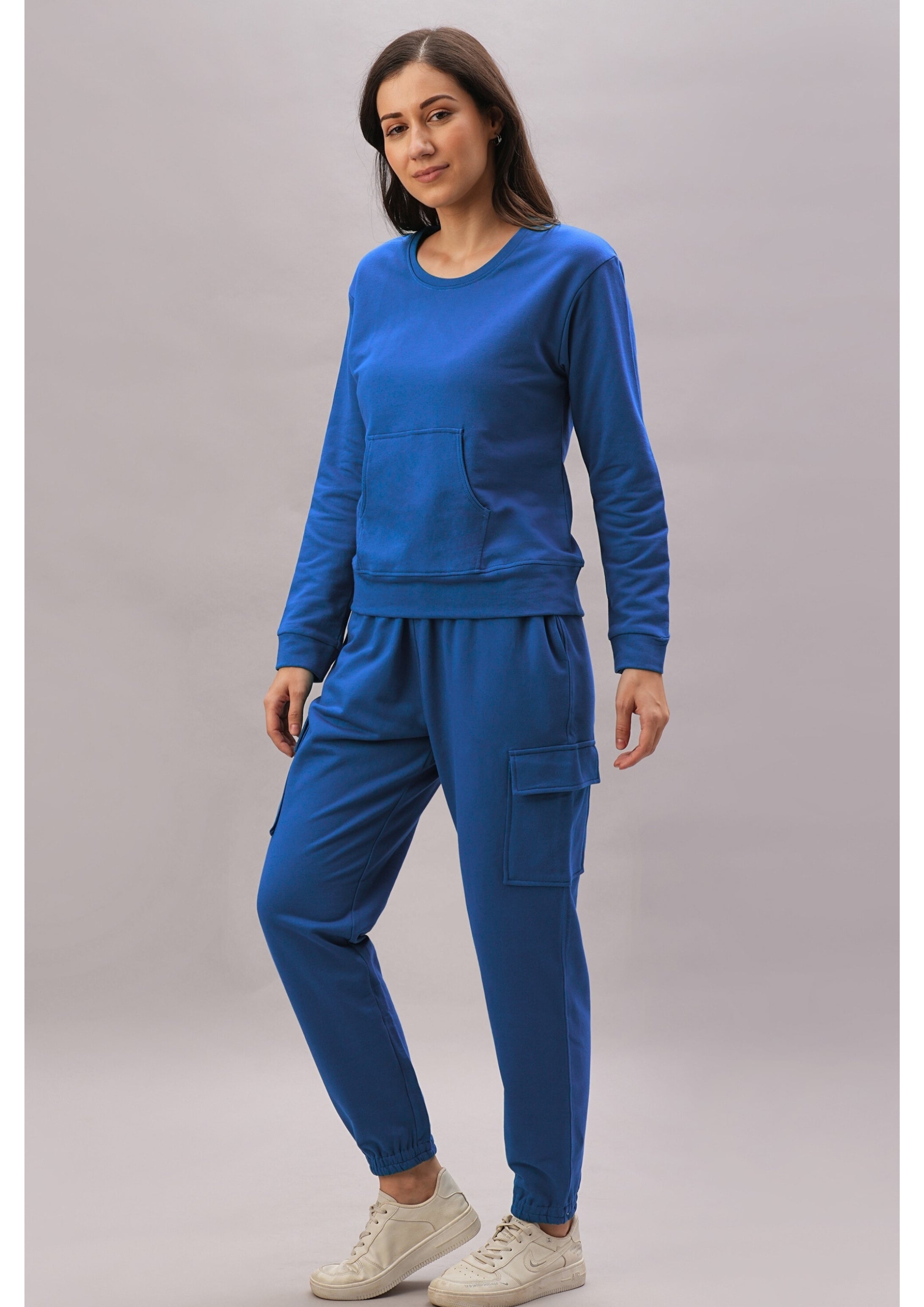 A woman stands against a plain background in the Soft Royal Blue Trackset, featuring cargo-style joggers and white sneakers, crafted from a breathable cotton-viscose blend.