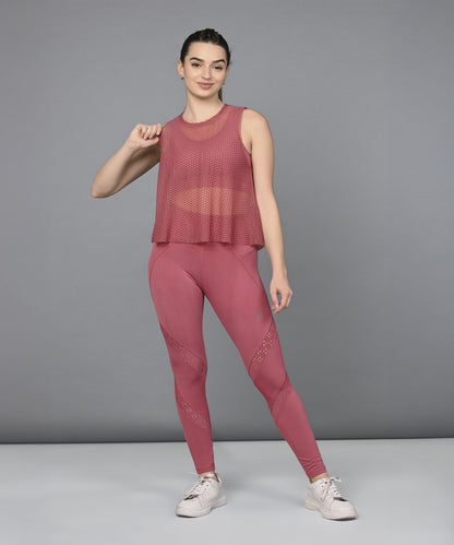 A woman poses confidently against a gray background, showcasing the Peony Pink Netted Top paired with white sneakers for a chic, sporty look.