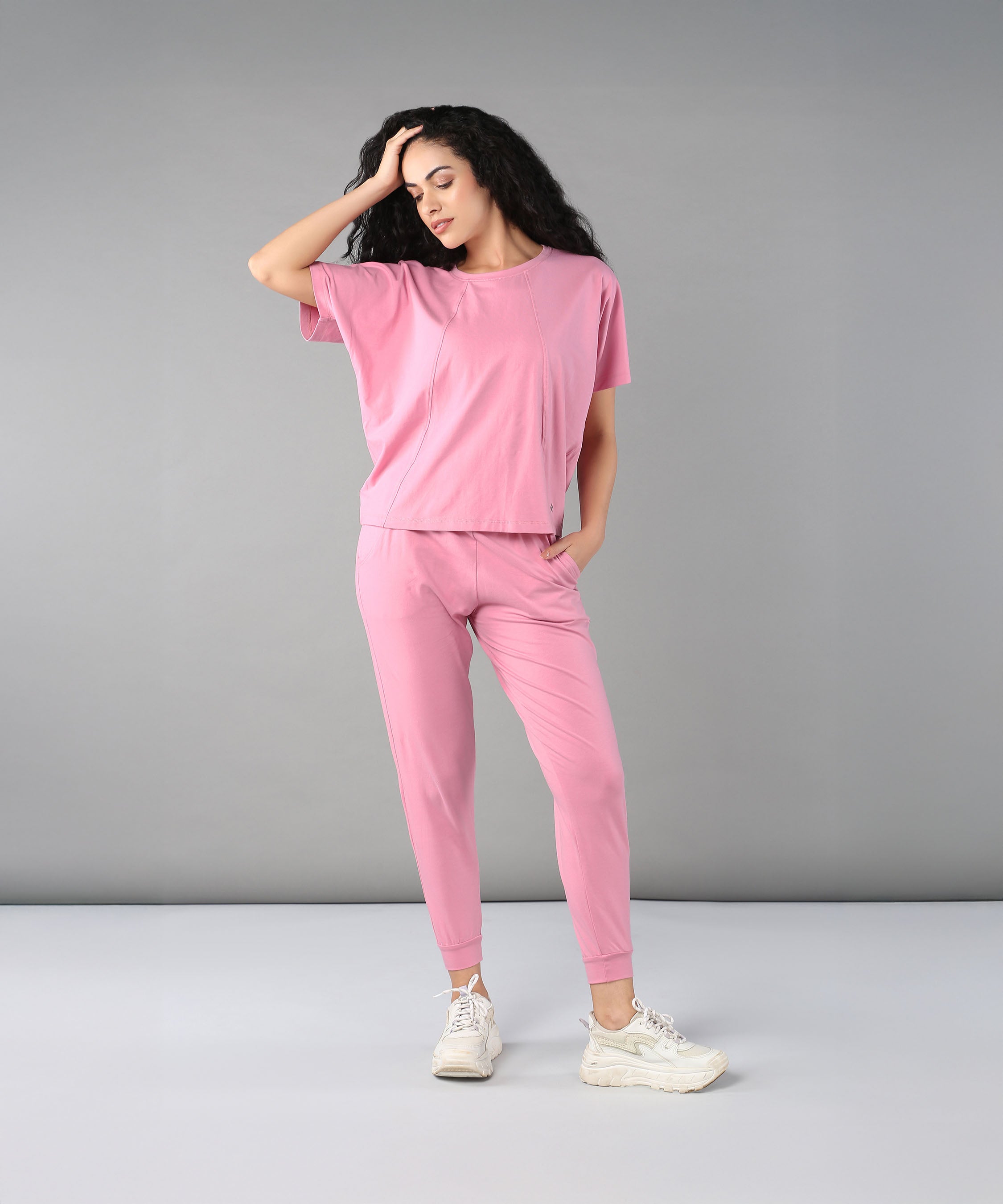 A woman wears the Flamingo Pink Chic Ensemble— a buttery soft Racer Back top and Joggers in cotton lycra—posing with one hand on her head against a plain grey background.