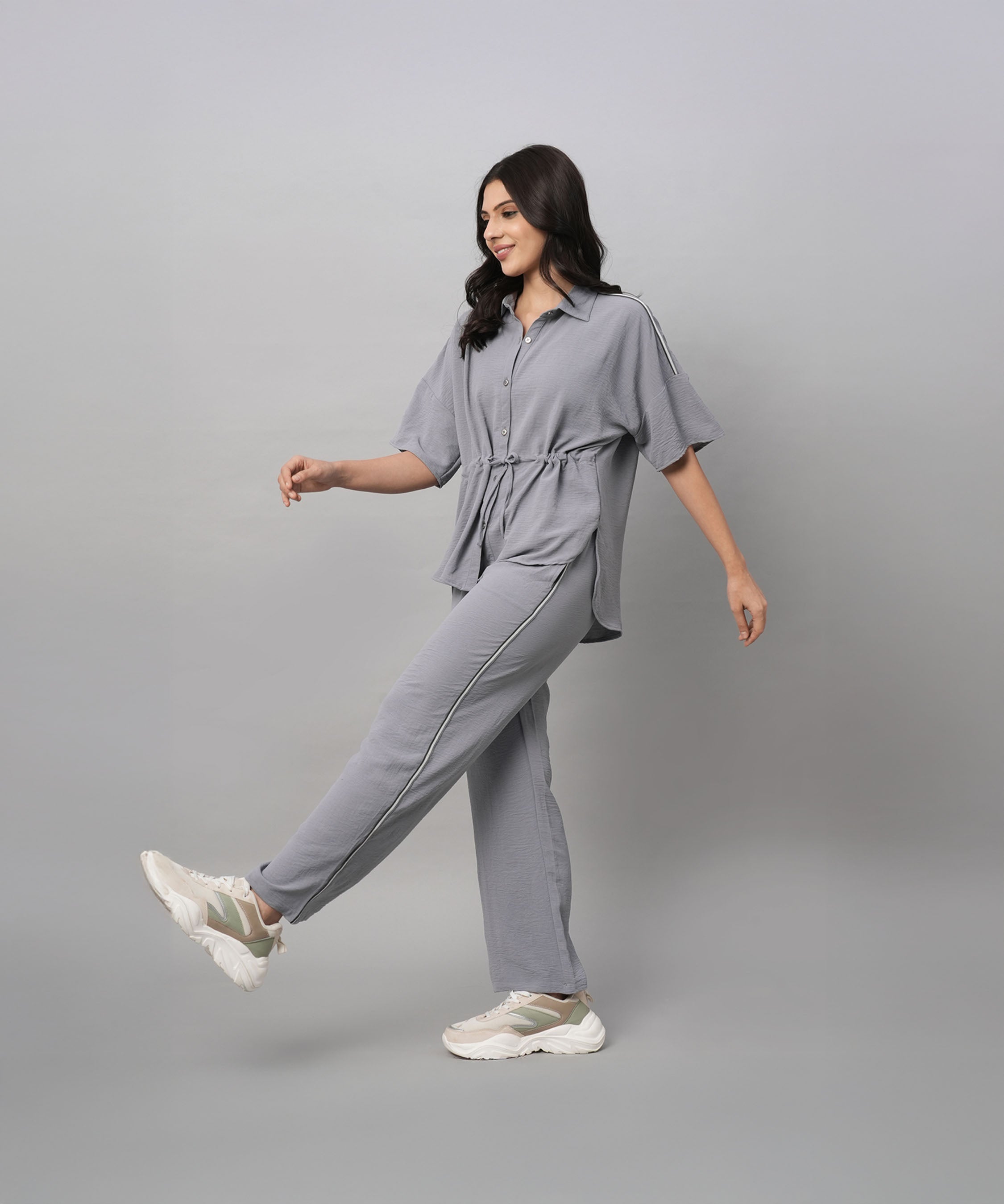 A woman wears the Grey Tie Up Top with Loose Pants Cord Set, pairing the coordinated grey outfit with white sneakers as she walks against a plain grey background.