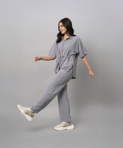 A woman wears the Grey Tie Up Top with Loose Pants Cord Set, pairing the coordinated grey outfit with white sneakers as she walks against a plain grey background.