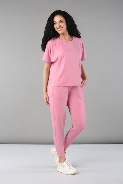 Woman in a Baby Pink Co-Ord Set featuring a cotton lycra t-shirt and elastic waistband trousers, smiling and standing against a gray background.