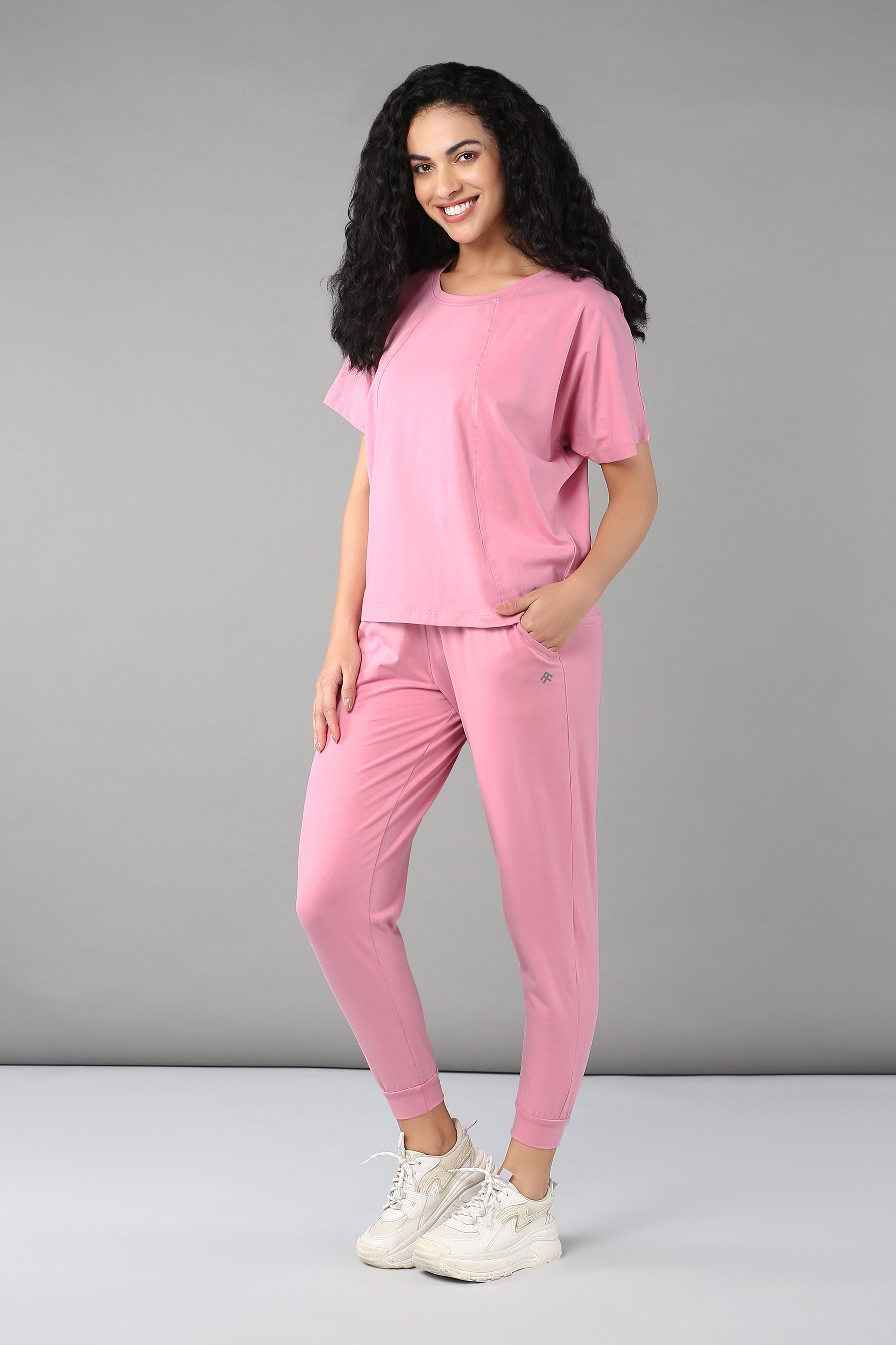 Woman with curly hair wears the Baby Pink Co-Ord Set, featuring a cotton lycra t-shirt and matching elastic waistband trousers, as she stands smiling against a gray background.