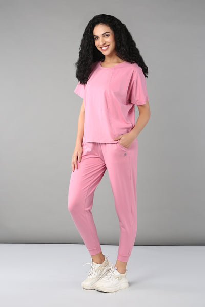 Woman with curly hair wears the Baby Pink Co-Ord Set, featuring a cotton lycra t-shirt and matching elastic waistband trousers, as she stands smiling against a gray background.