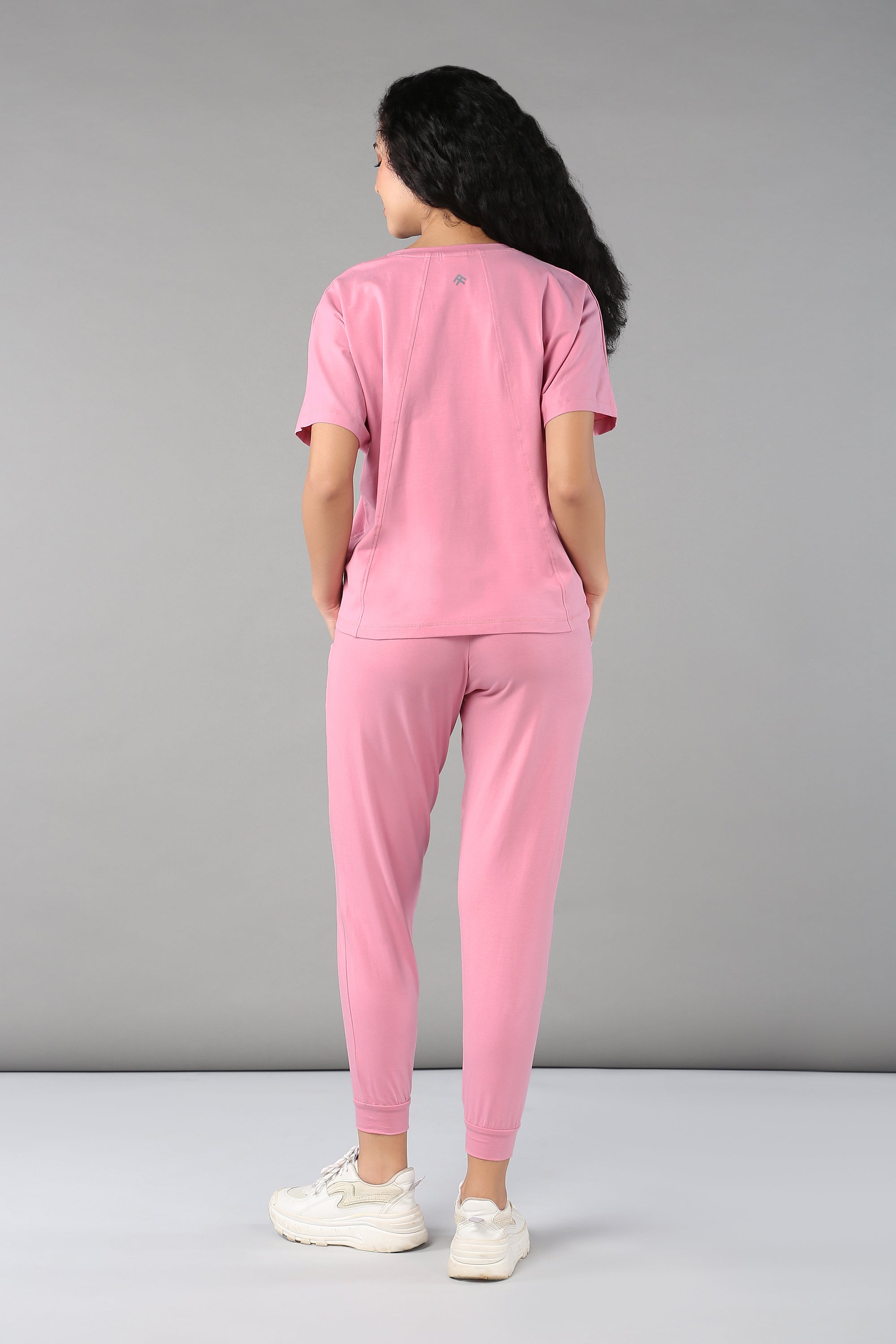 A woman faces away, wearing the Baby Pink Co-Ord Set featuring breathable fabric, elastic waistband trousers, and white sneakers against a gray background.