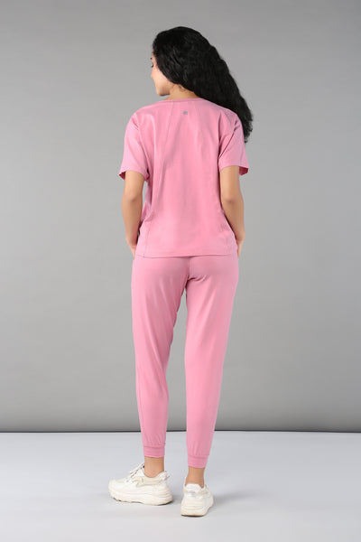 A woman faces away, wearing the Baby Pink Co-Ord Set featuring breathable fabric, elastic waistband trousers, and white sneakers against a gray background.
