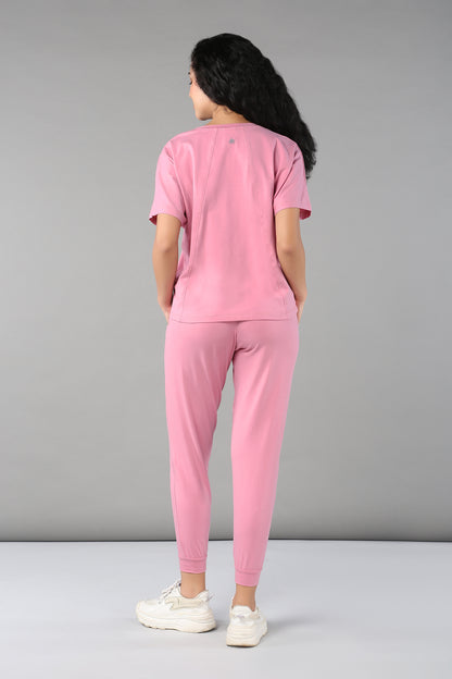 A woman faces away, wearing the Baby Pink Co-Ord Set featuring breathable fabric, elastic waistband trousers, and white sneakers against a gray background.