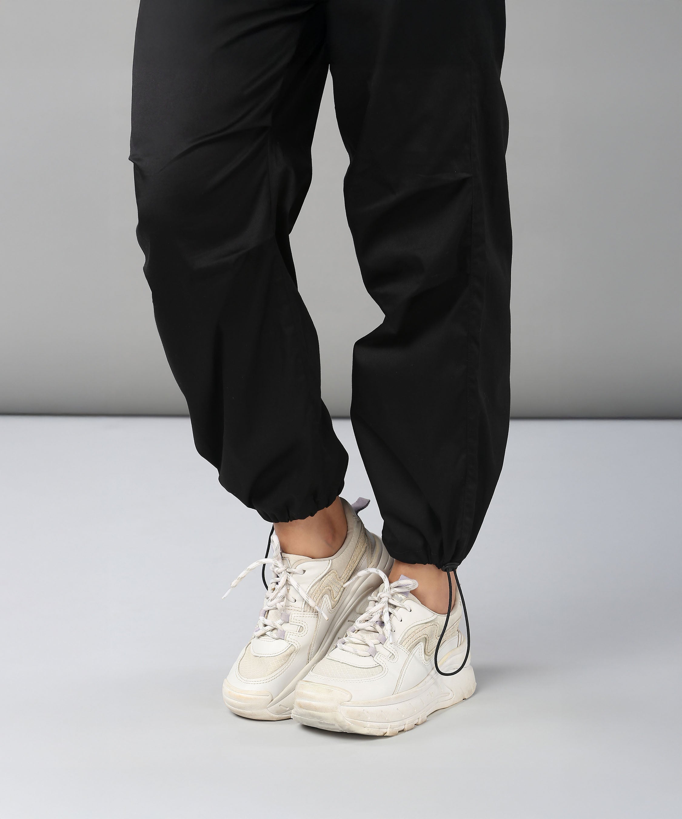 A person wearing Black Parachute Pants stands on a light grey floor with legs slightly crossed, paired with white sneakers.