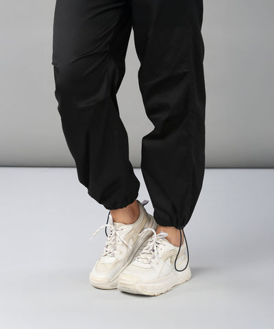 A person wearing Black Parachute Pants stands on a light grey floor with legs slightly crossed, paired with white sneakers.
