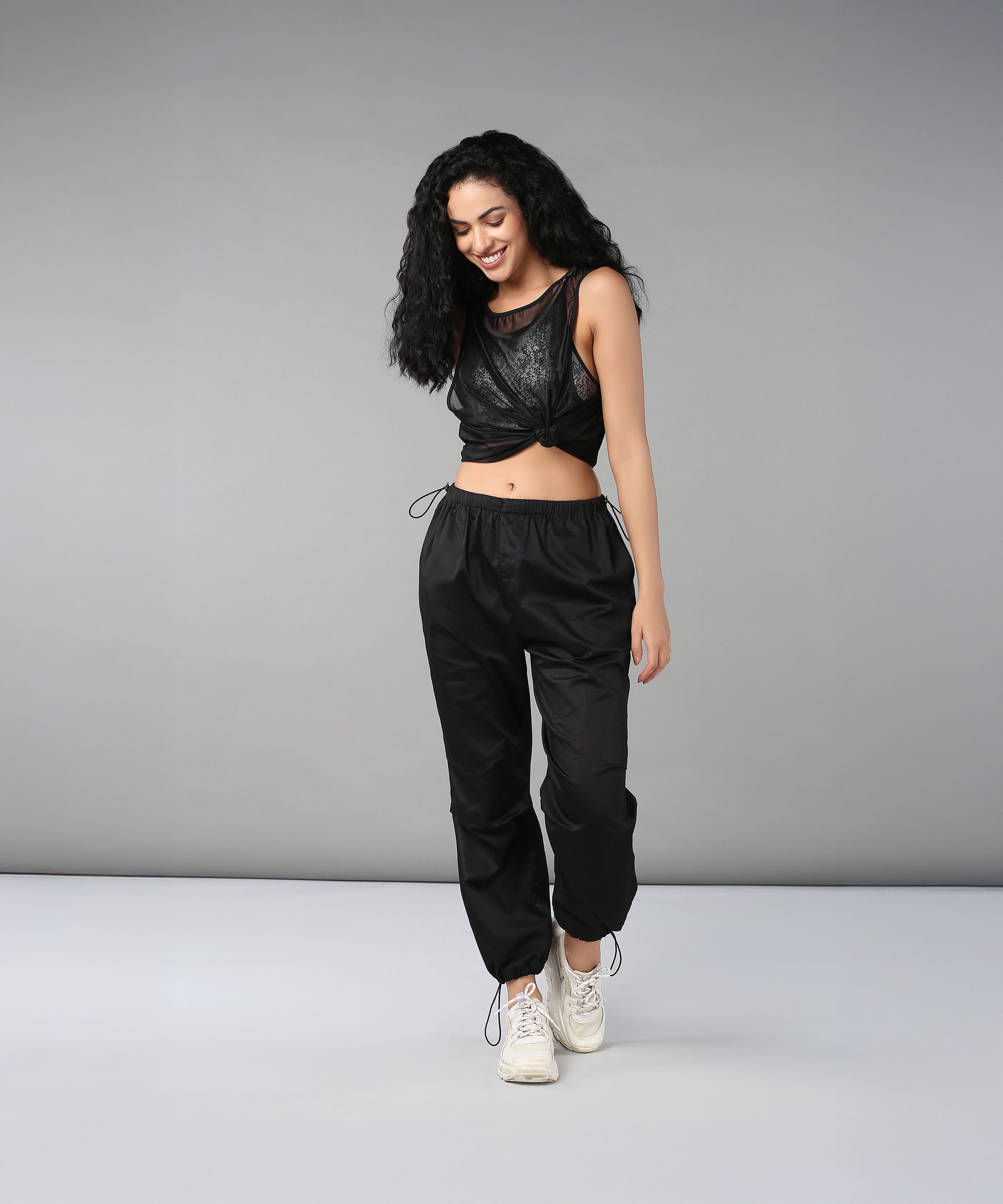 Woman with curly hair stands, smiling, in black adjustable waist Black Parachute Pants, a black crop top, and white sneakers.