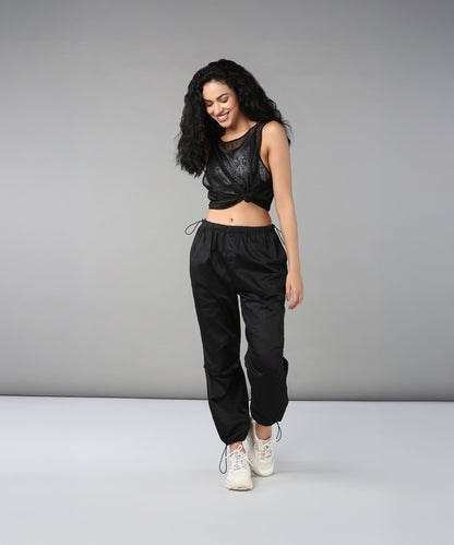 Woman with curly hair stands, smiling, in black adjustable waist Black Parachute Pants, a black crop top, and white sneakers.