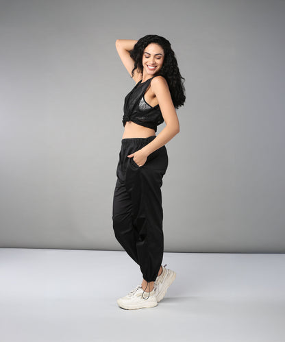A smiling woman stands sideways against a gray background, posing with her hand on her head while wearing Black Parachute Pants and a crop top.