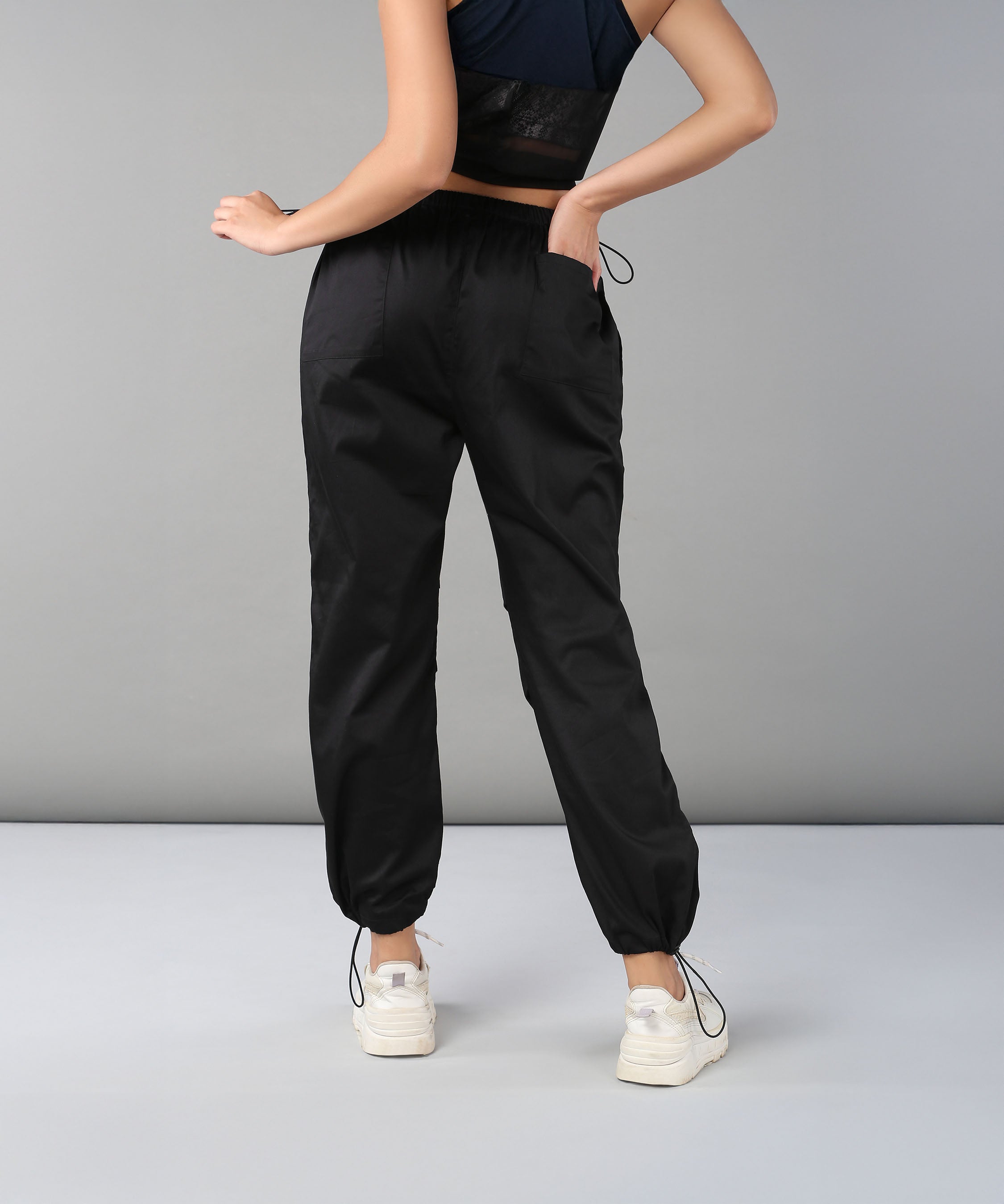 A woman wearing Black Parachute Pants and white sneakers stands with her back to the camera against a plain gray background.