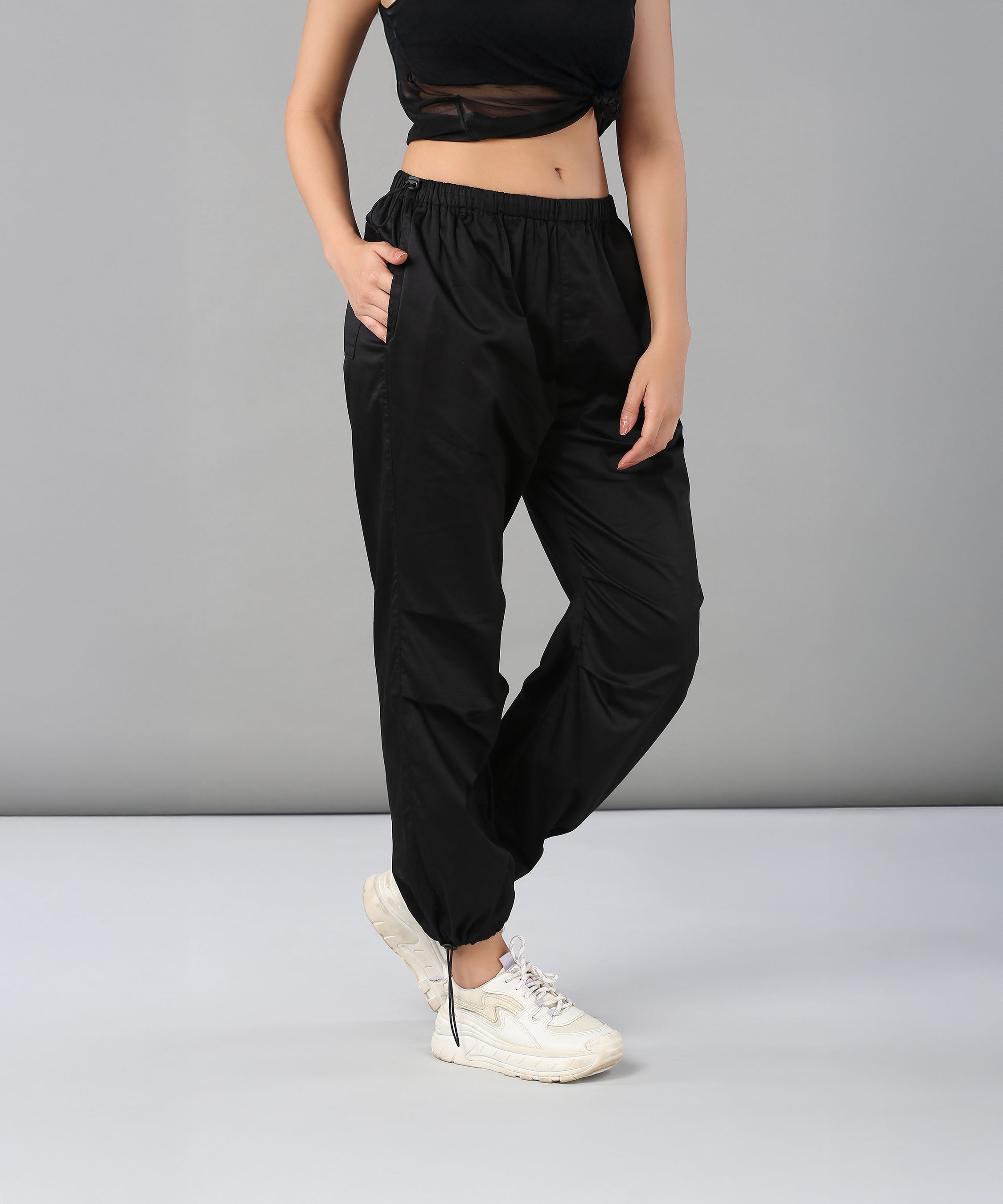 Woman wearing Black Parachute Pants with an adjustable waist and white sneakers poses against a plain gray background.