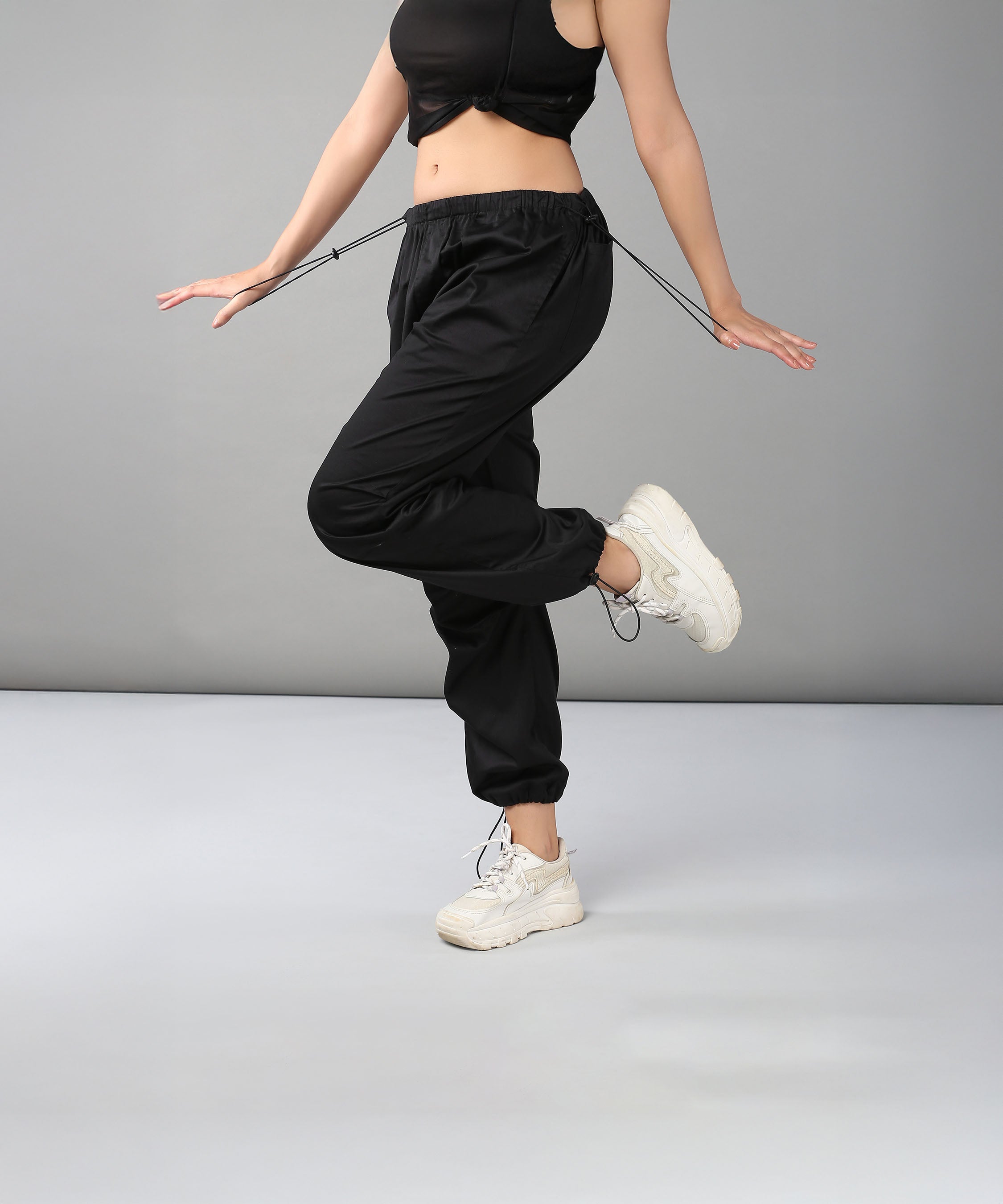 A woman in Black Parachute Pants and white sneakers balances on one leg against a plain gray background.