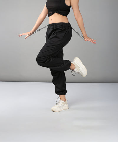 A woman in Black Parachute Pants and white sneakers balances on one leg against a plain gray background.