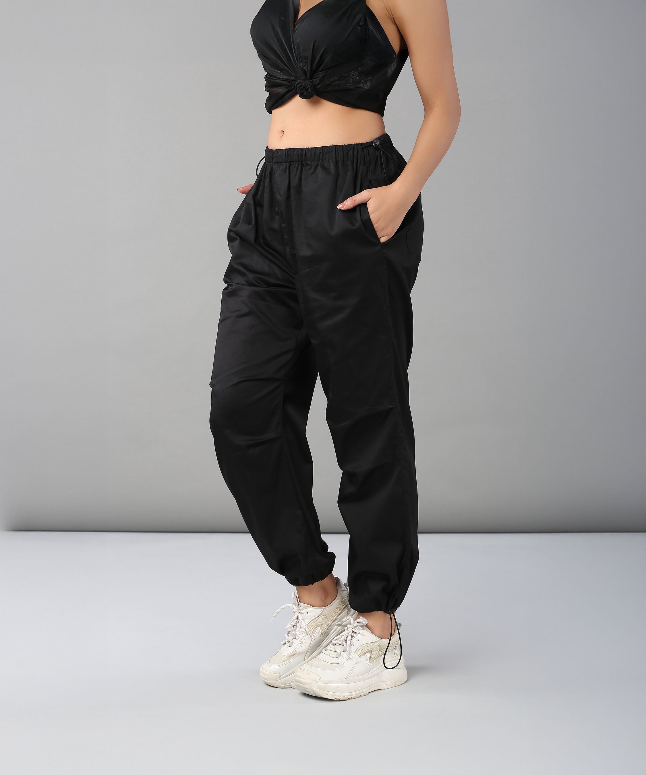 Woman in Black Parachute Pants with an adjustable waist, paired with a black crop top and white sneakers, standing against a gray background.