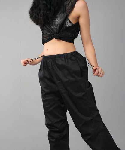 Woman wearing Black Parachute Pants with an adjustable waist, pulling the drawstrings, paired with a sleeveless black top against a plain background.