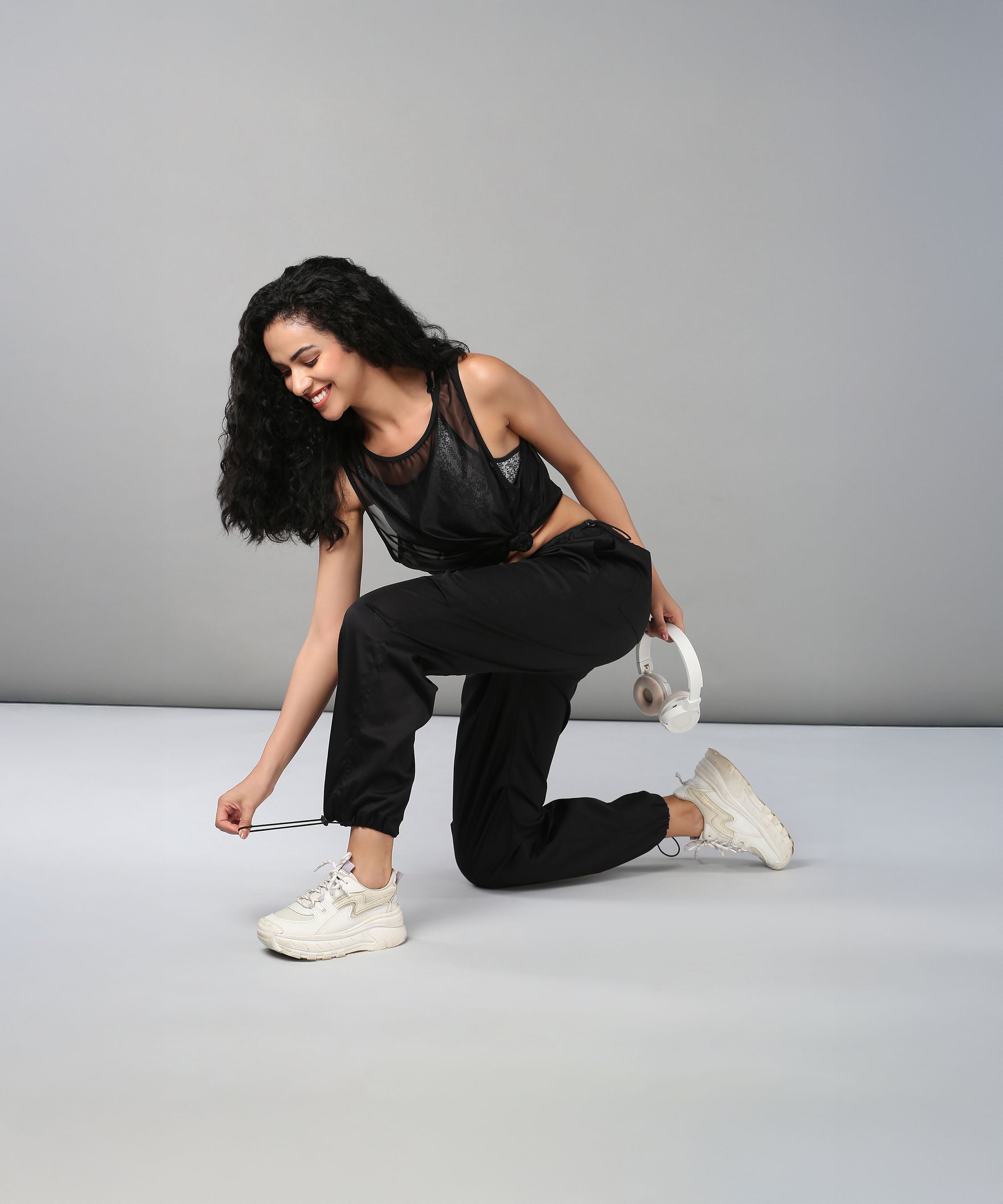 A woman wearing Black Parachute Pants kneels to tie her shoe, smiling with white headphones in one hand.