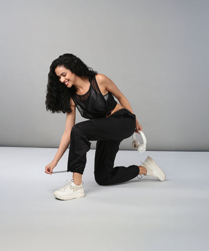 A woman wearing Black Parachute Pants kneels to tie her shoe, smiling with white headphones in one hand.