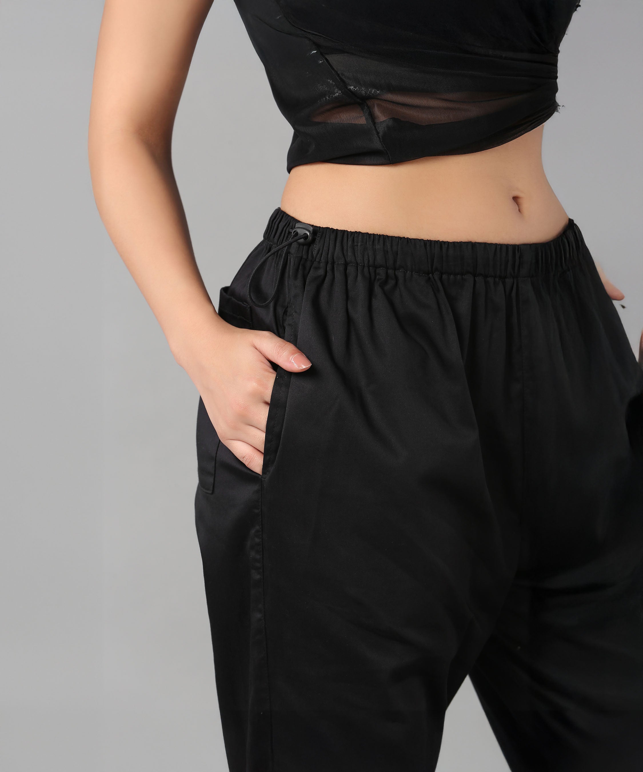 Model wears Black Parachute Pants and a black crop top, hand in pocket, shown from shoulders to mid-thigh.