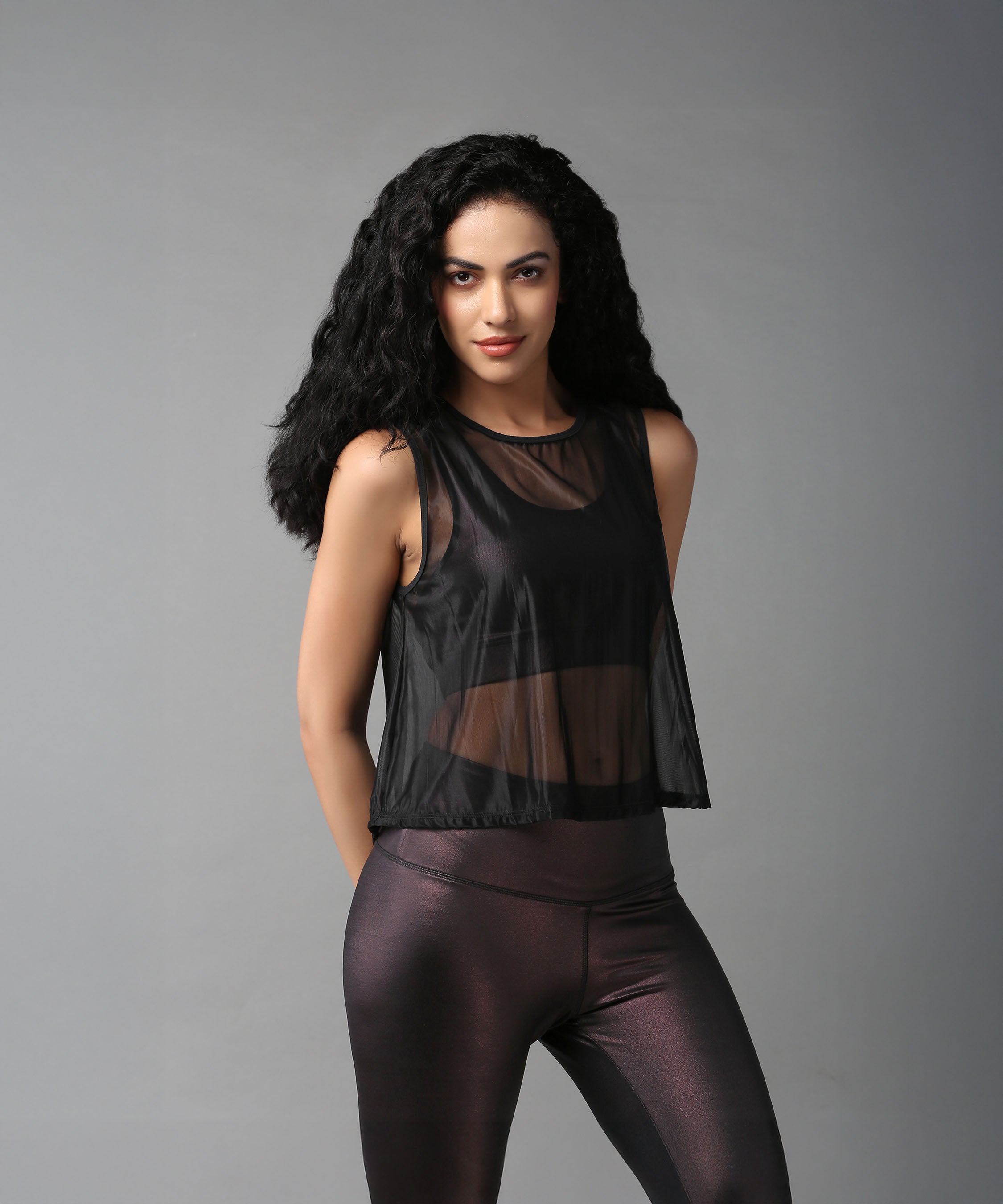 A woman with long curly hair models the Black Sheer Top and shiny leggings, posing against a plain gray background.