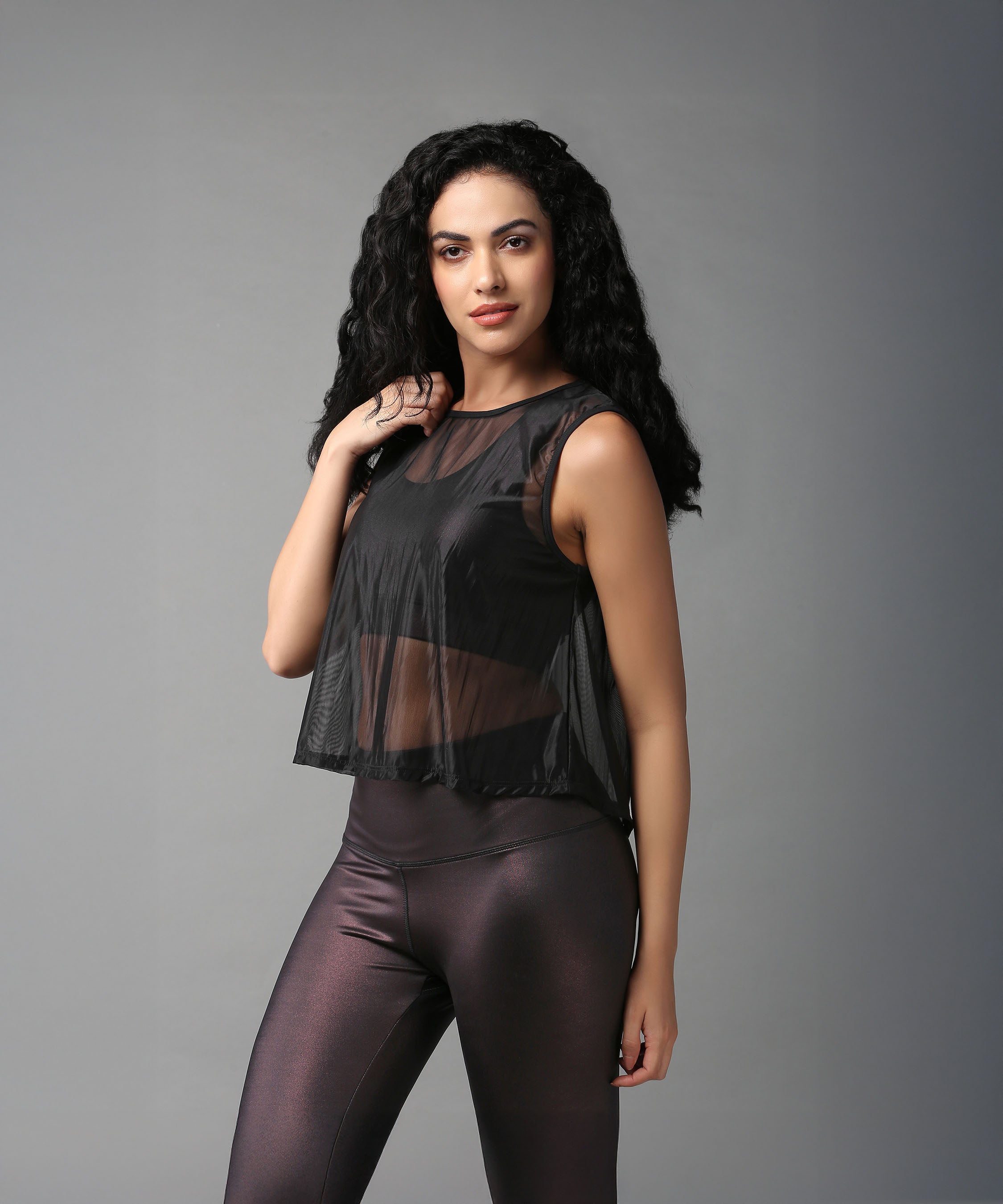 Woman with long curly hair poses confidently in a Black Sheer Top and shiny black leggings.