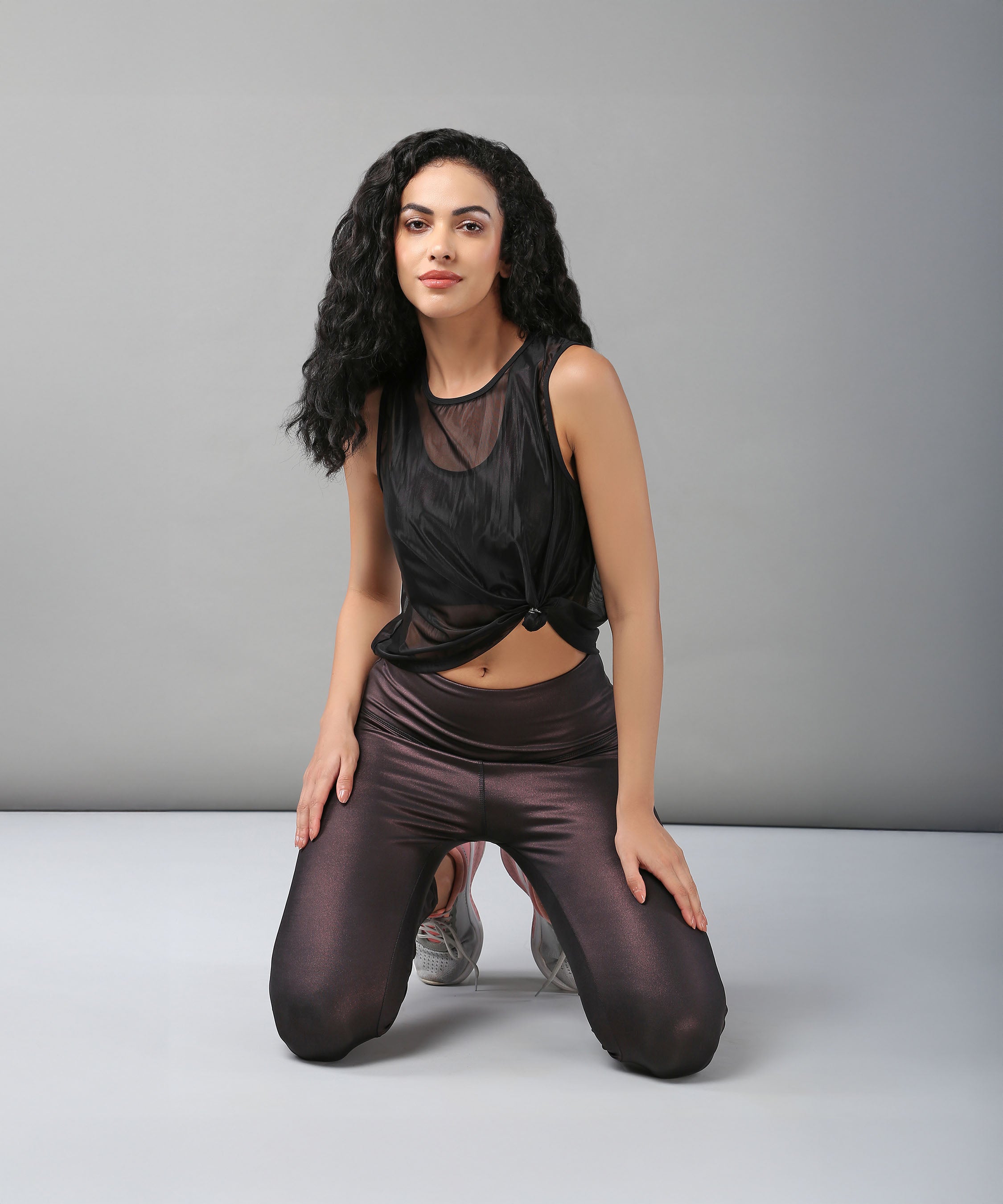 Woman with long curly hair kneeling on the floor, wearing a sexy Black Sheer Top and shiny dark leggings.