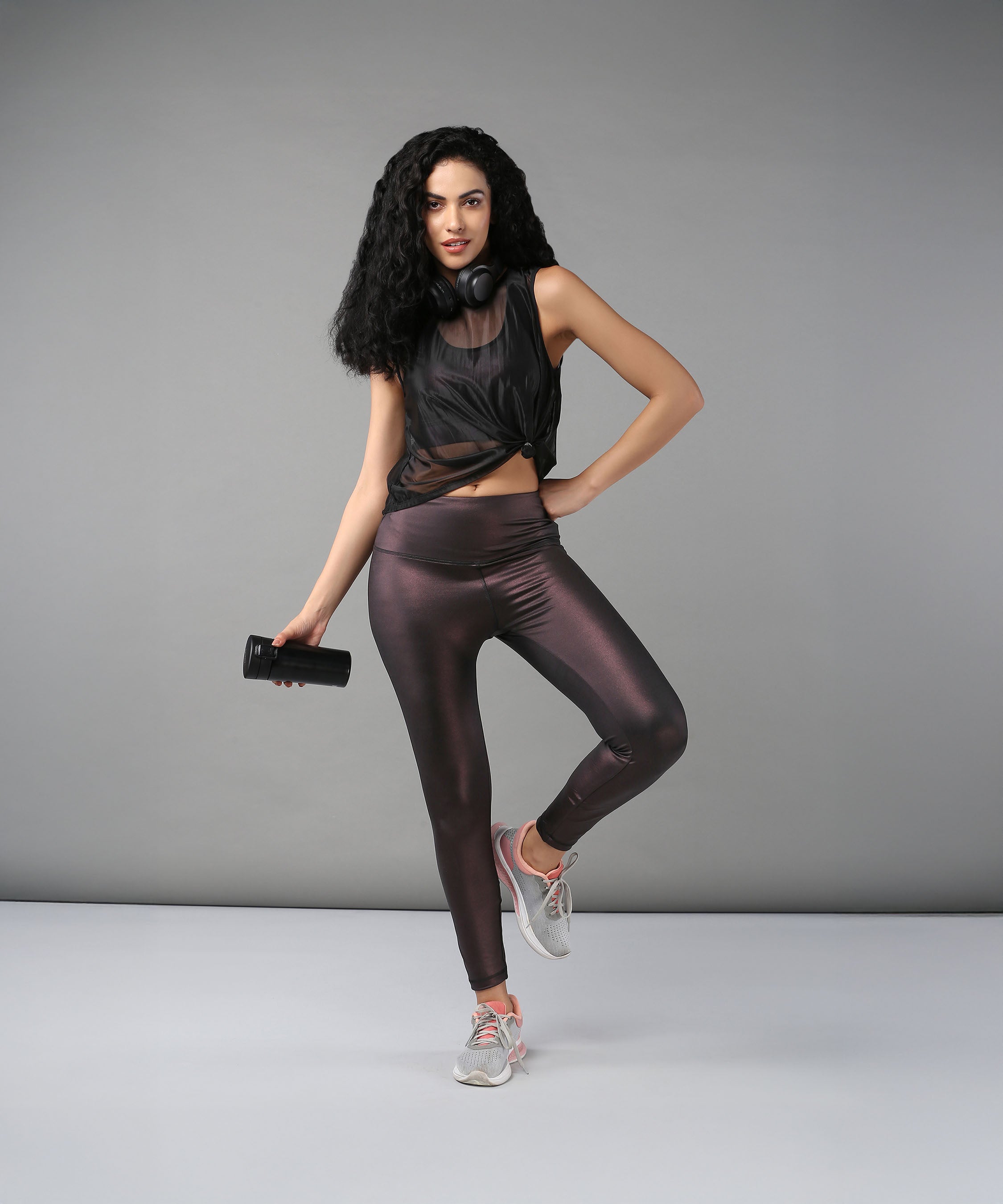 Confident woman poses against a gray background wearing headphones, a Black Sheer Top, shiny workout clothes, and holding a water bottle.