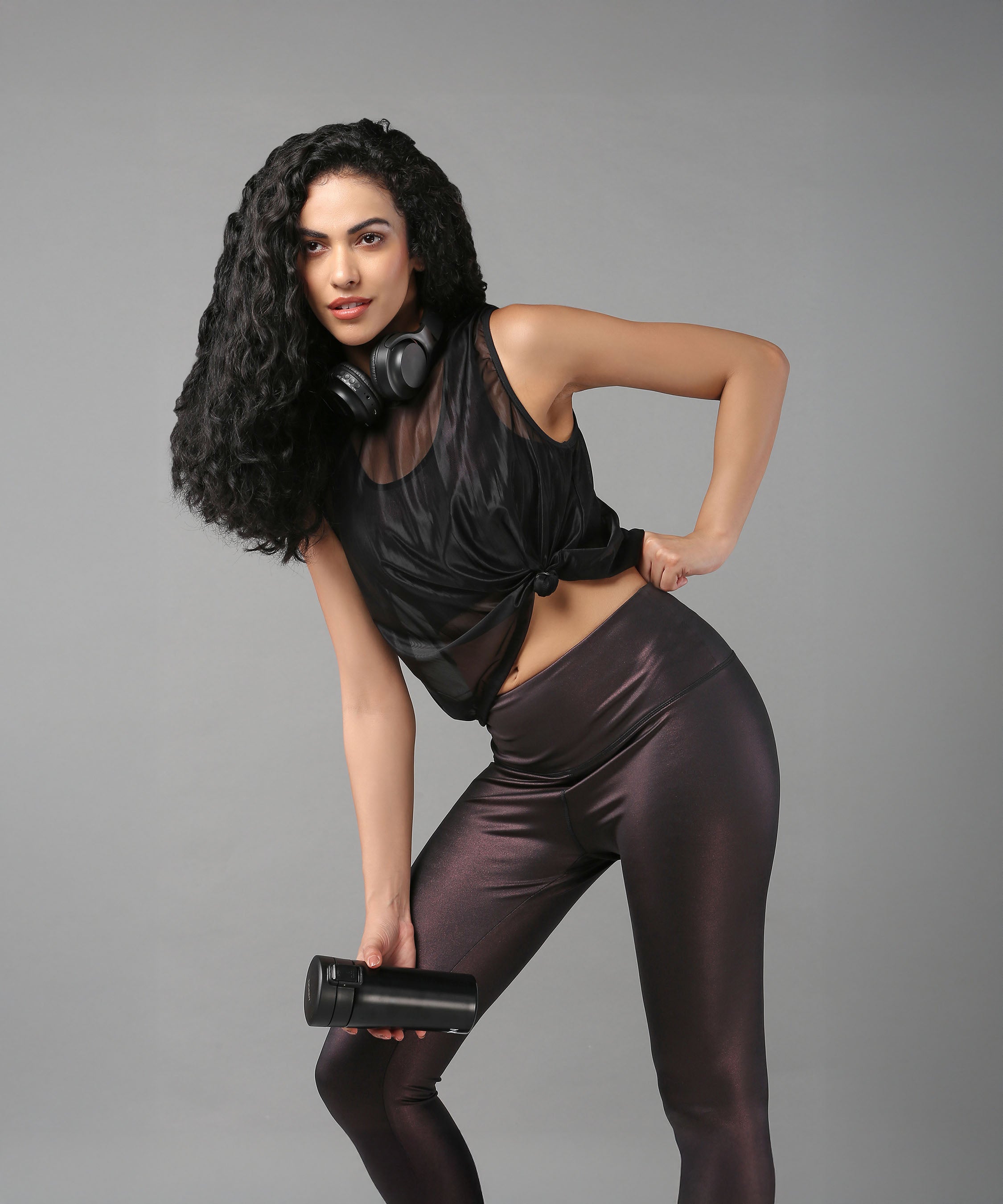 A woman wearing the Black Sheer Top poses with headphones around her neck and a water bottle in hand, set against a gray background.